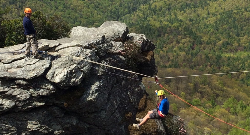 A person wearing safety gear is secured by ropes as they execute a Tyrolean traverse above a wooded area 